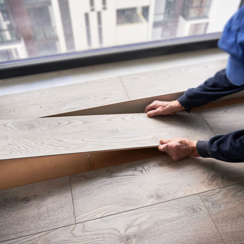 Male builder installing laminate flooring in room with large window. Man construction worker laying laminate planks on the floor in apartment under renovation. Hardwood floor renovation concept.